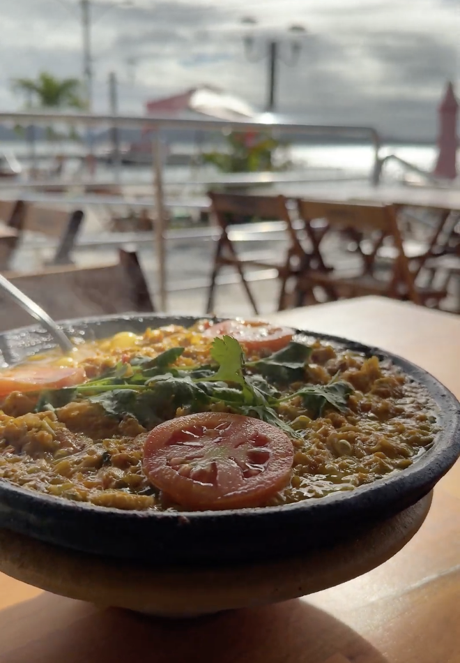 Prato típico baiano em panela de barro com tomates e coentro, restaurante à beira-mar ao fundo