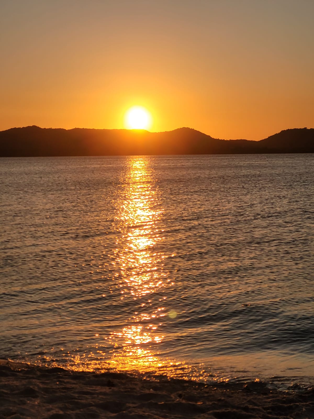 Pôr do sol em Salinas da Margarida — sol laranja se pondo atrás das colinas com reflexo dourado no mar