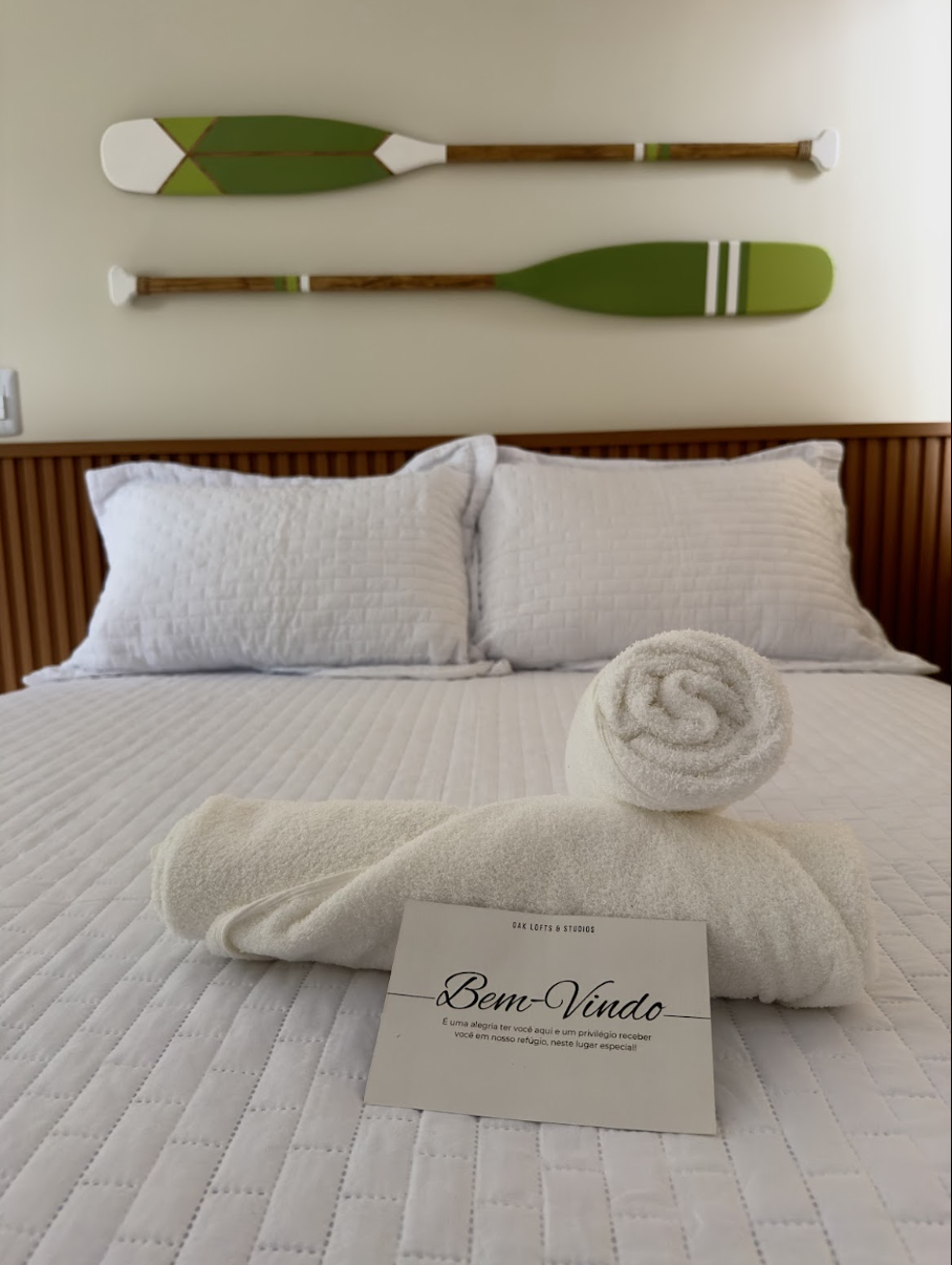 Cama branca com toalha enrolada e cartão Bem-Vindo da OAK, remos decorativos verdes na parede e cabeceira de ripado em madeira
