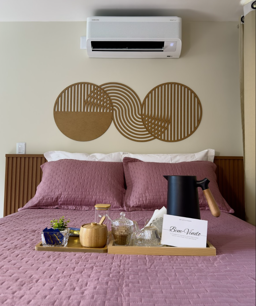 Cama com roupa de cama rosê, arte dourada de círculos na parede e bandeja Bem-Vindo com chaleira preta