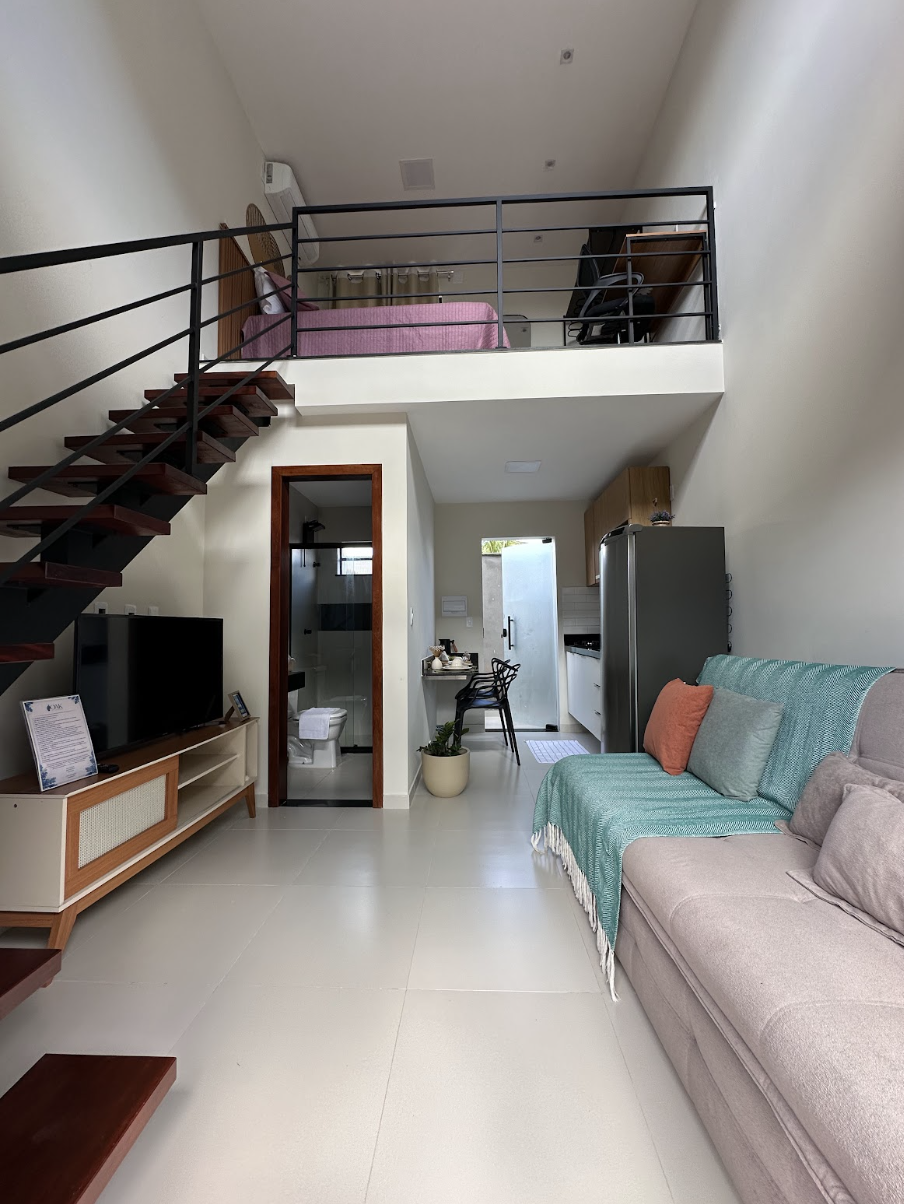 Sala do loft duplex com sofá, TV e escada para o mezanino com cama rosê ao fundo