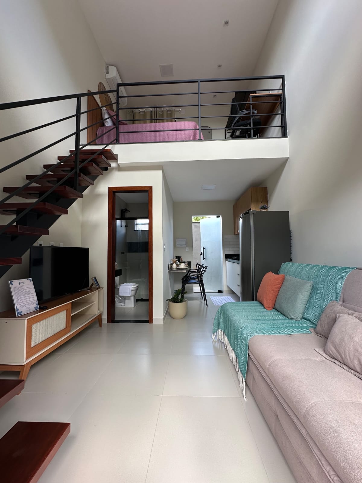 Vista ampla da sala do loft duplex com sofá bege, escada de madeira e mezanino com cama rosê visível