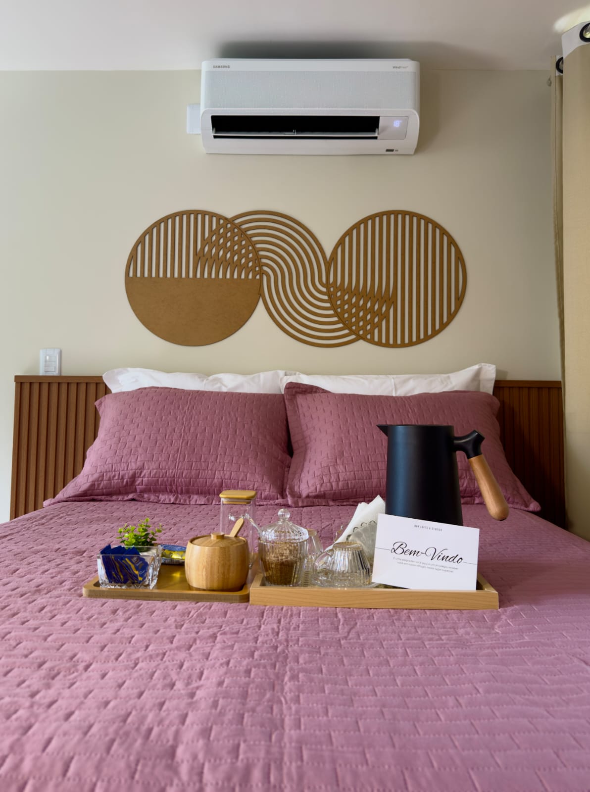 Cama rosê com bandeja Bem-Vindo, chaleira preta e arte dourada de círculos na parede